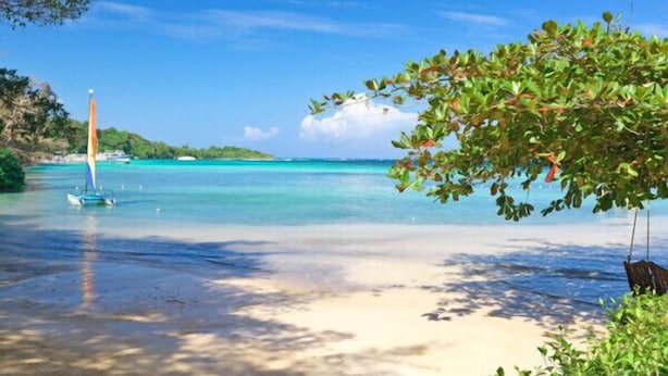 Een strand met helder turquoise water, een zeilboot en een hangmat onder een boom.