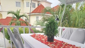 A covered outdoor dining area with a white table and chairs, a potted plant, and a red and gold table runner. - PinkHibiscus Guest House (Montego Bay)