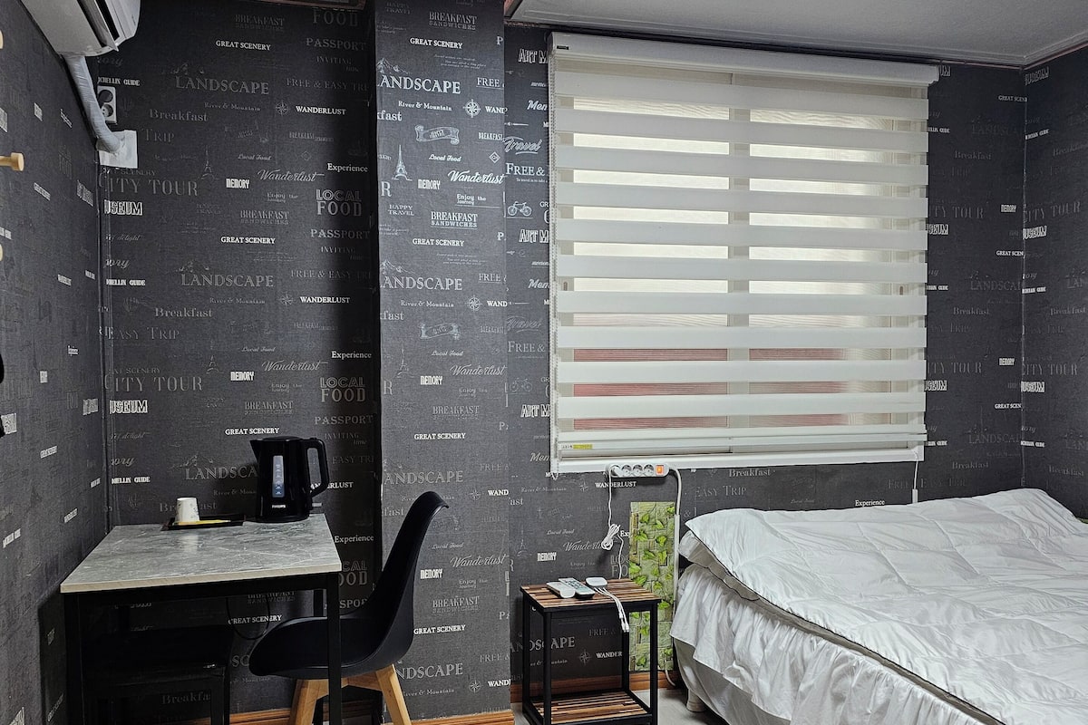 Ein Schlafzimmer mit einem Bett, einem Schreibtisch mit Stuhl, einem Fenster mit Jalousien und einer Wand mit aufgedrucktem Text.