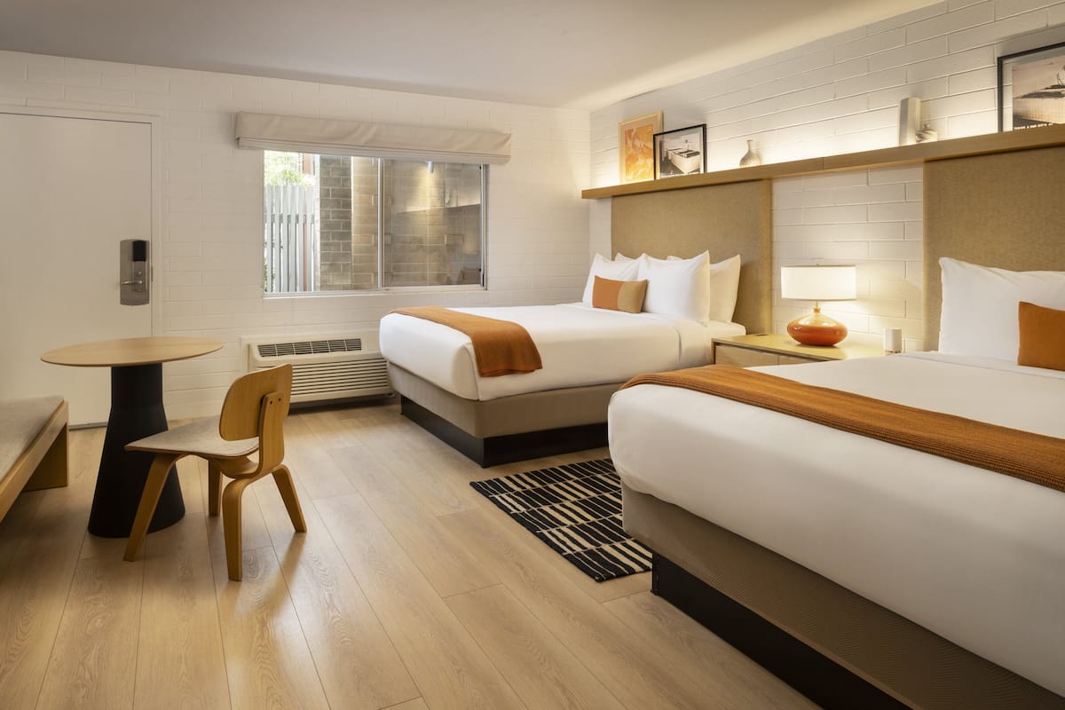 A hotel room with two beds, a table, a chair, a heater, and a window with blinds.