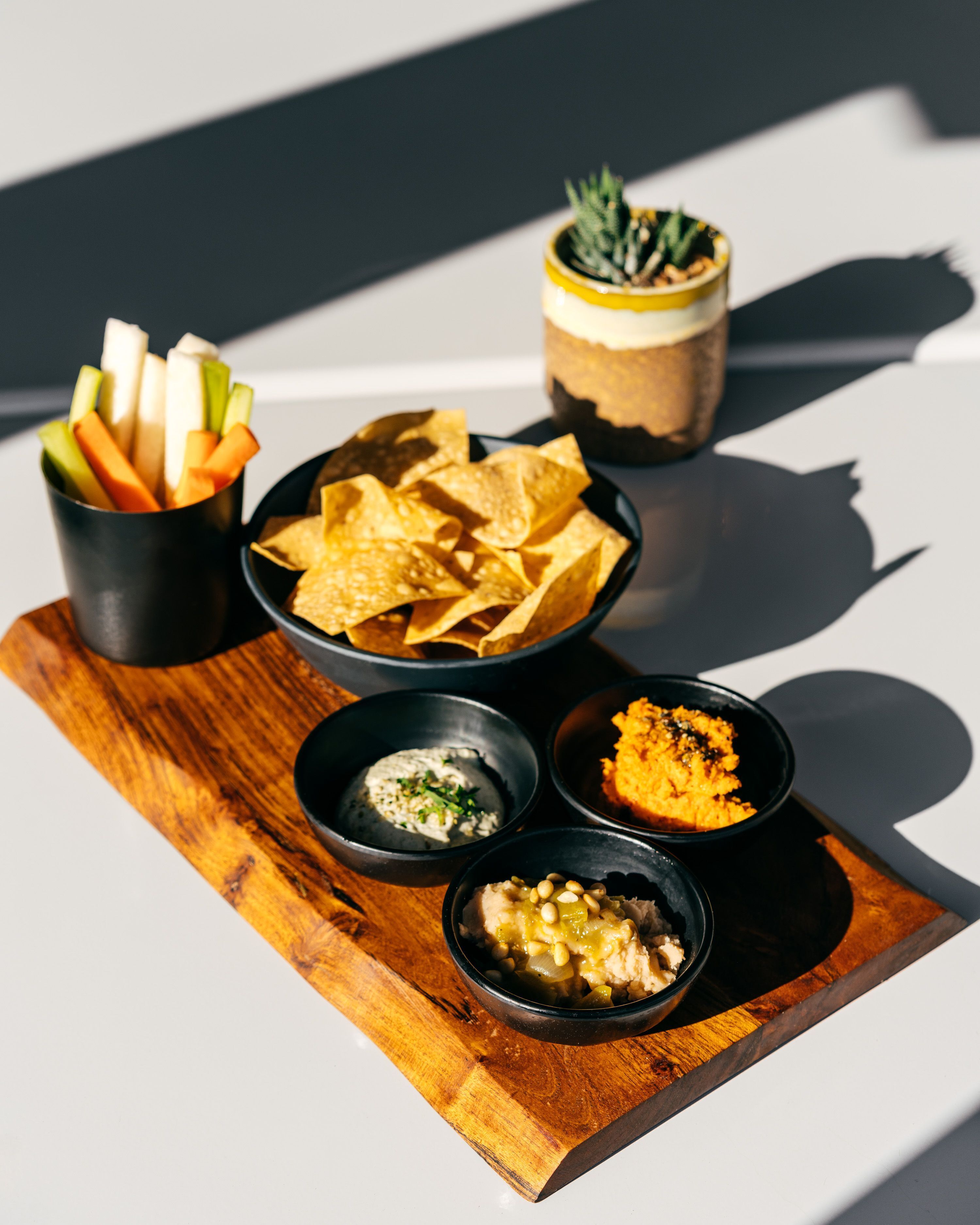 A wooden board with various food items including chips, dips, and a small potted plant.