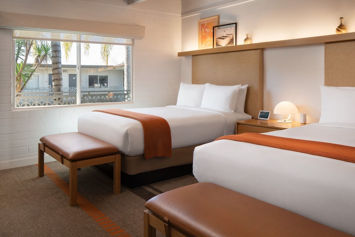 A hotel room with two beds, a bench, a nightstand, and a window with a view of palm trees.