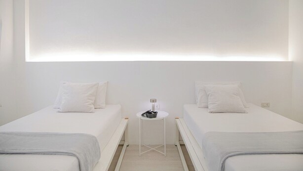 A minimalist hotel room with two beds, a small table, and a wall-mounted light fixture. - HAO HOSTEL PHUKET (Phuket)