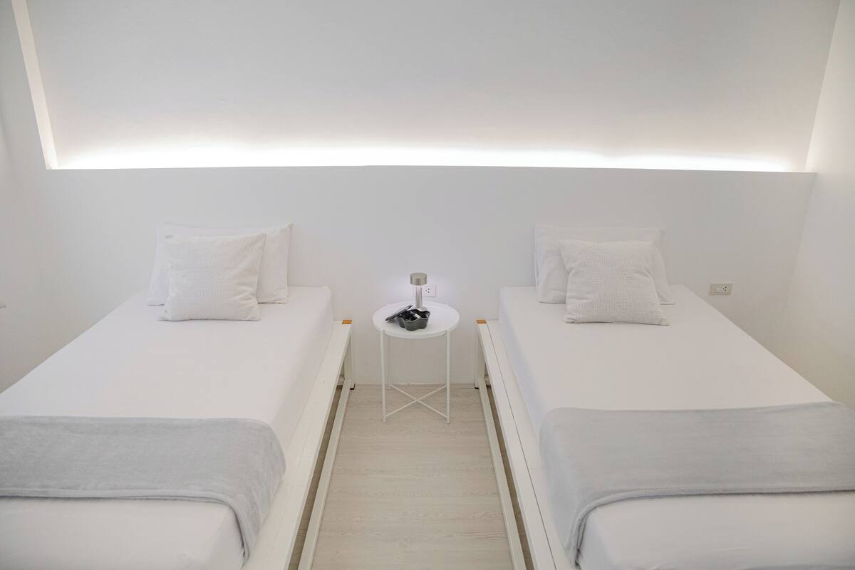 A small, modern room with two single beds, a small table with a lamp, and a ceiling light.