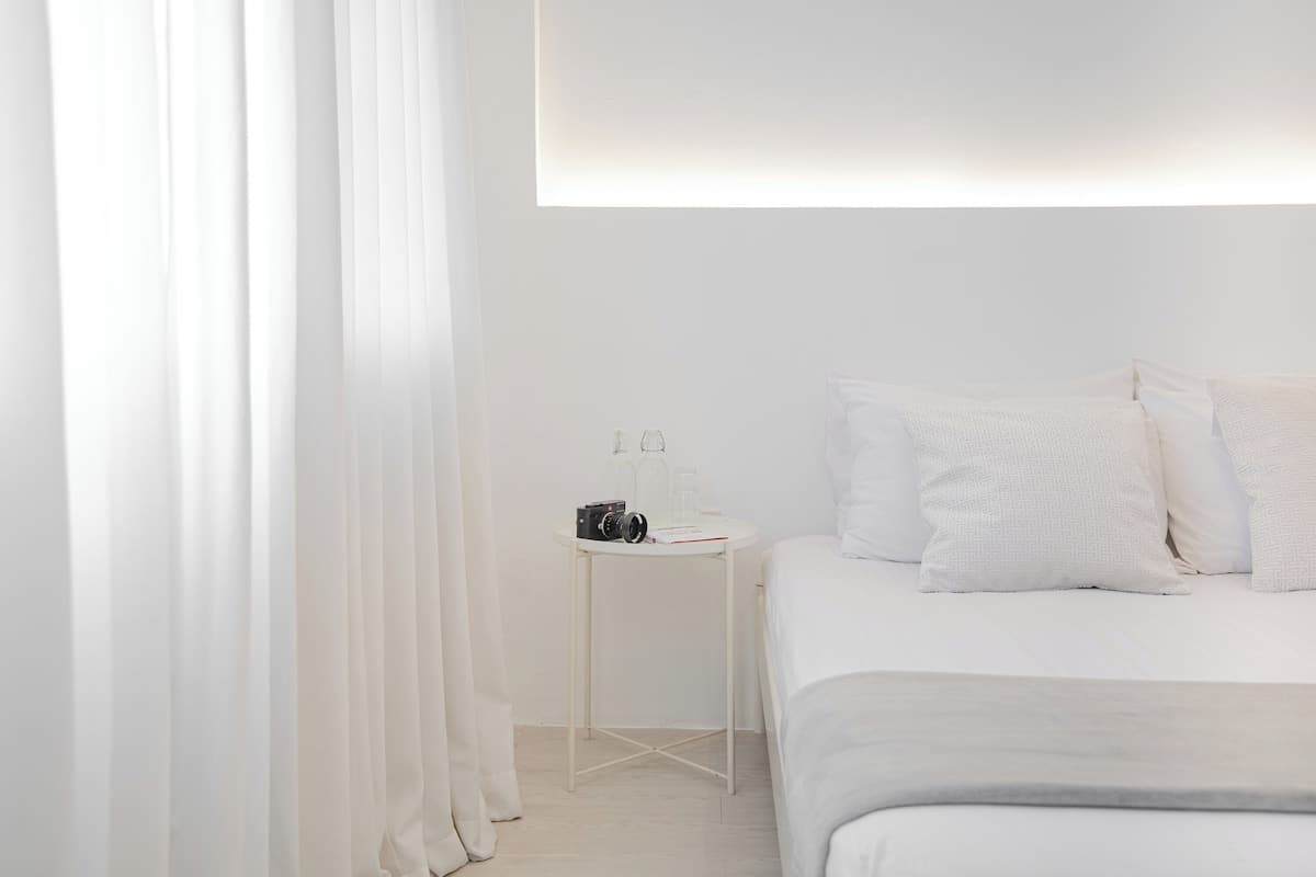 A minimalist bedroom with a neatly made bed, a small bedside table with a camera and glasses, and a white wall.