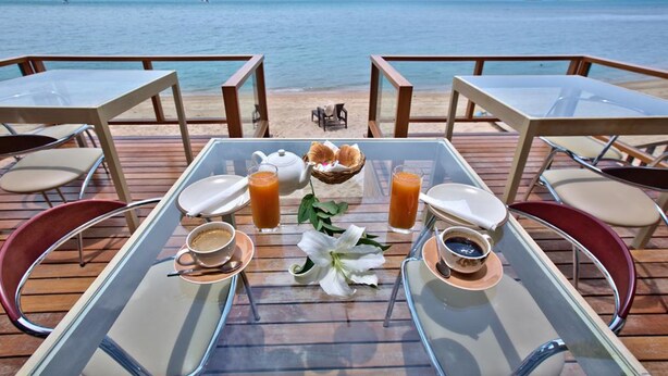 Una mesa preparada con alimentos para el desayuno y una flor, con vista a una playa y barcos a lo lejos.
