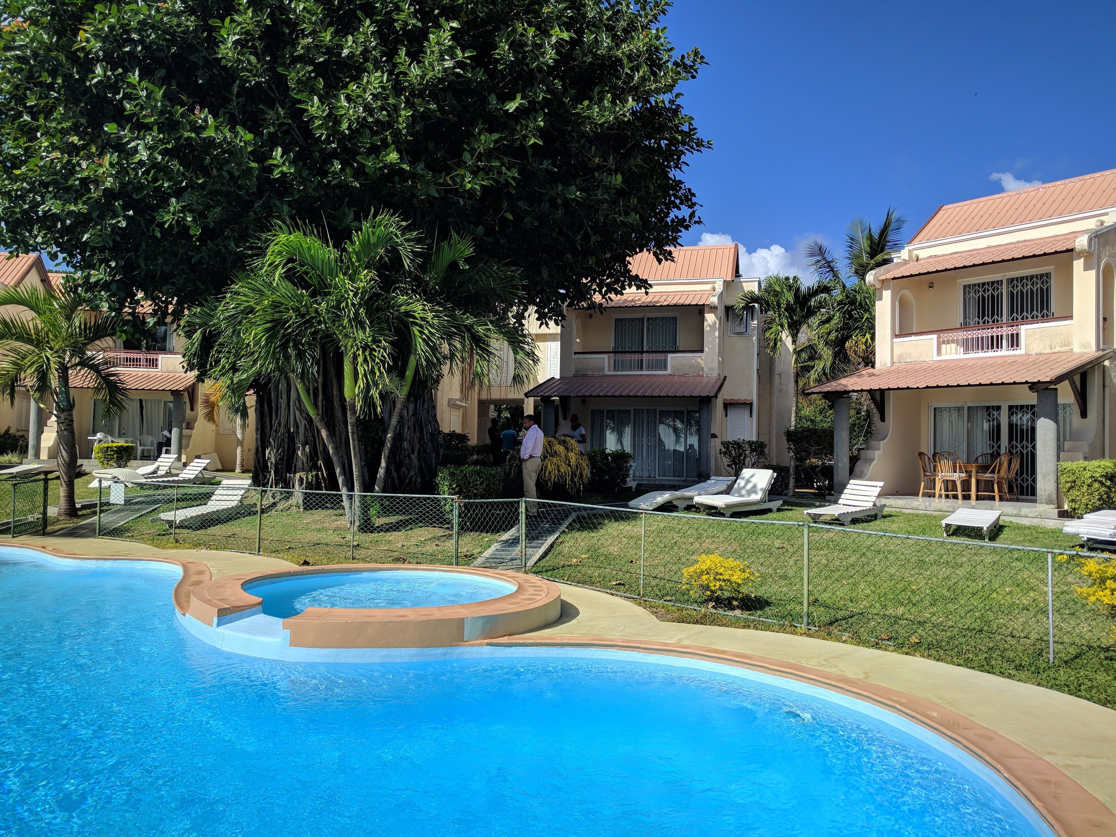 Una piscina con sillas de descanso y un área cercada, rodeada de palmeras y un edificio con tejado de azulejos.