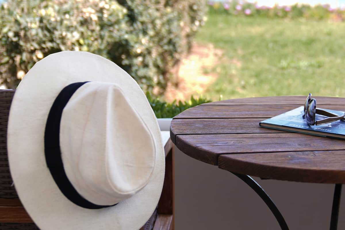 A white hat on a wicker chair, a notebook with a pen, and a pair of glasses on a wooden table with a view of the sea and greenery.