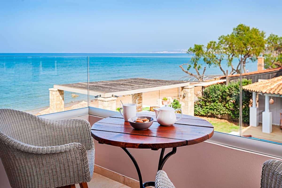 A balcony with wicker chairs, a wooden table with a bowl and teapots, and a view of the sea and buildings.