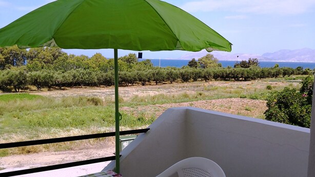 A balcony with a table and chair, a green umbrella, and a view of a vineyard and sea. - Yiannis Apartments (Kos)