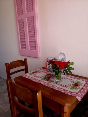 Una mesa de madera con mantel floral, un jarrón con flores rojas y un folleto de alquiler de coches en la pared.