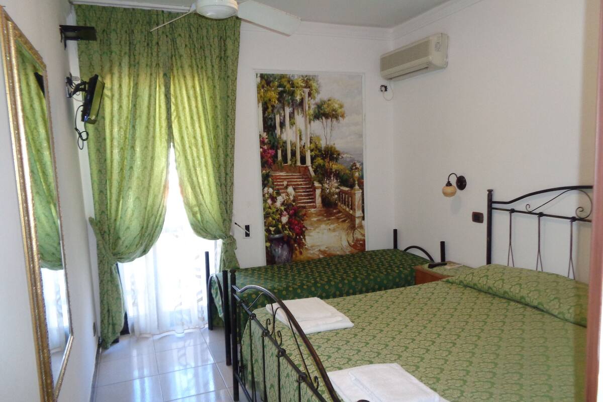 Ein Schlafzimmer mit Bett, Deckenventilator, Klimaanlage, einem Bild an der Wand und einem Fenster mit Vorhängen.