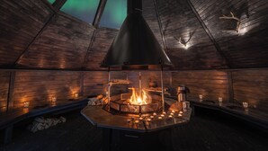 Un intérieur chaleureux en bois, avec une cheminée centrale, des bancs en bois et une vue sur les aurores boréales grâce à une verrière.