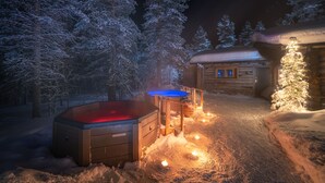 Une cabane en bois avec un bain à remous et une petite table, entourée d’arbres couverts de neige.