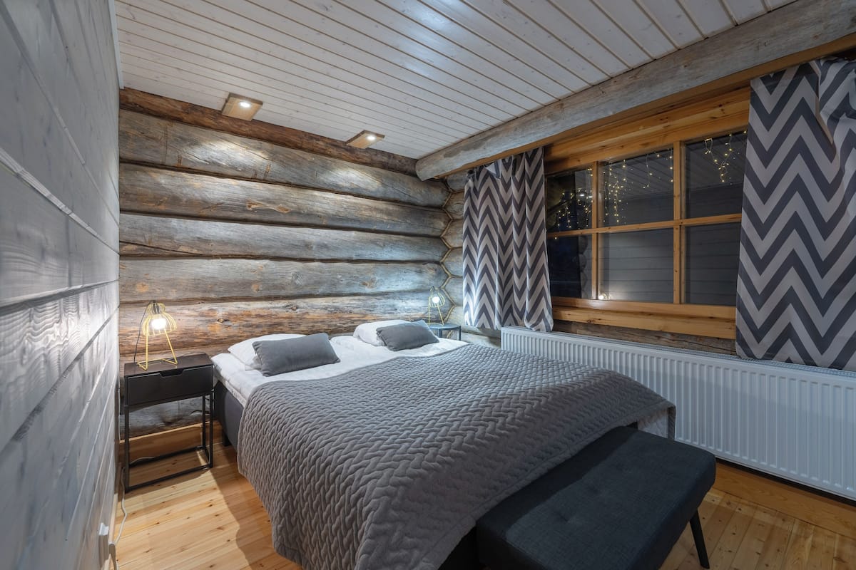 Une chambre à coucher située dans un chalet en rondins, avec un lit, une table de chevet, une lampe et une fenêtre avec des rideaux.
