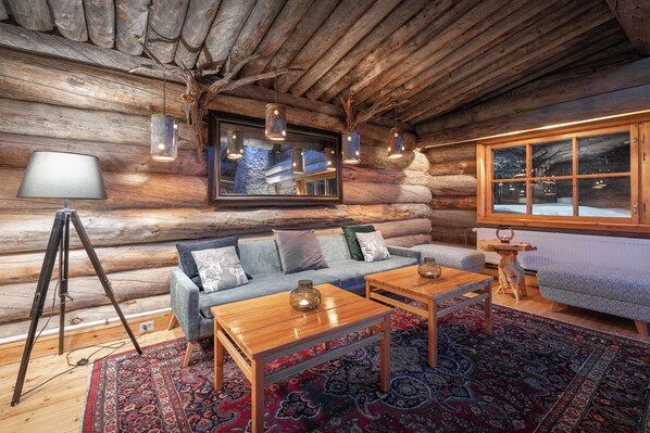 L’intérieur d’une cabane en rondins, avec un canapé, deux tables en bois, une lampe et une grande fenêtre.