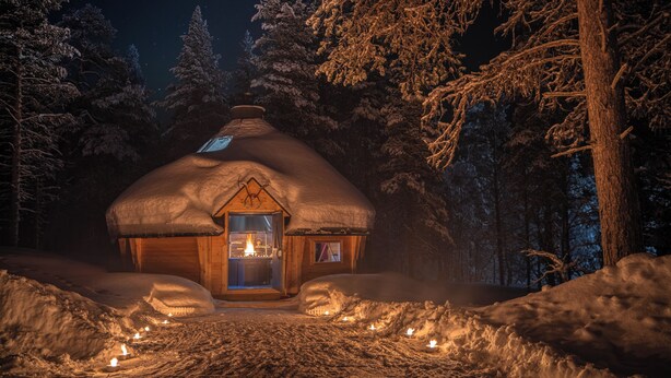 Une cabane confortable au toit de chaume, entourée d’arbres couverts de neige, dont la lumière chaleureuse se reflète à travers les fenêtres.