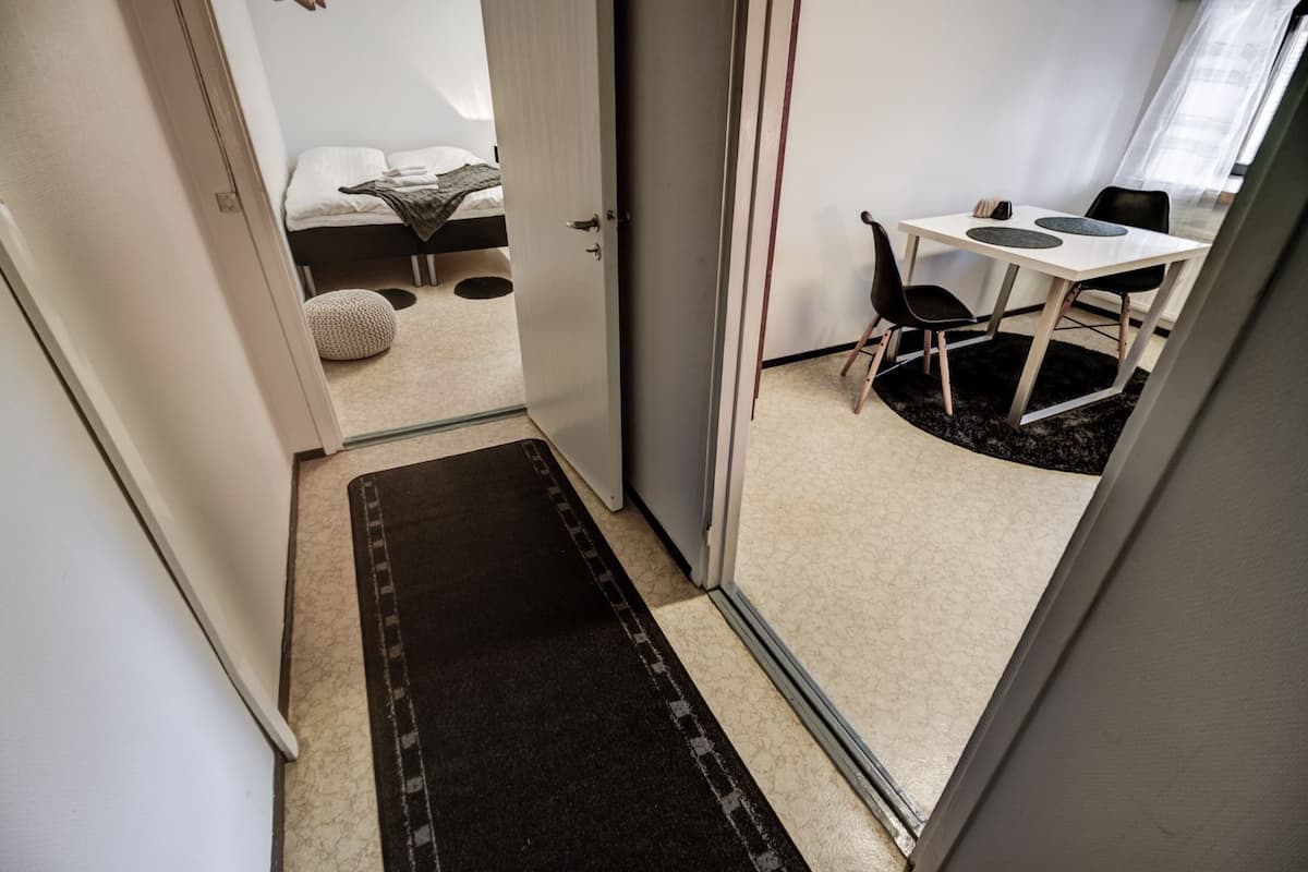 A narrow hallway leading to a bedroom with a bed, a small round rug, and a desk with chairs.