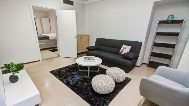 A modern living room with a grey sofa, a round coffee table, and two round poufs. A white side table with a plant is visible. -  Kuukkeli Apartments Sodankylä (Sodankyla)