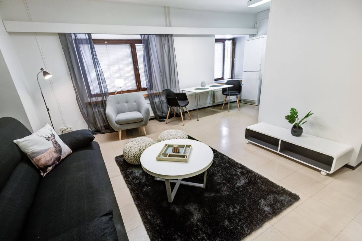 A modern living room with a black sofa, a round coffee table, and a grey armchair. There is a desk with two chairs and a potted plant.