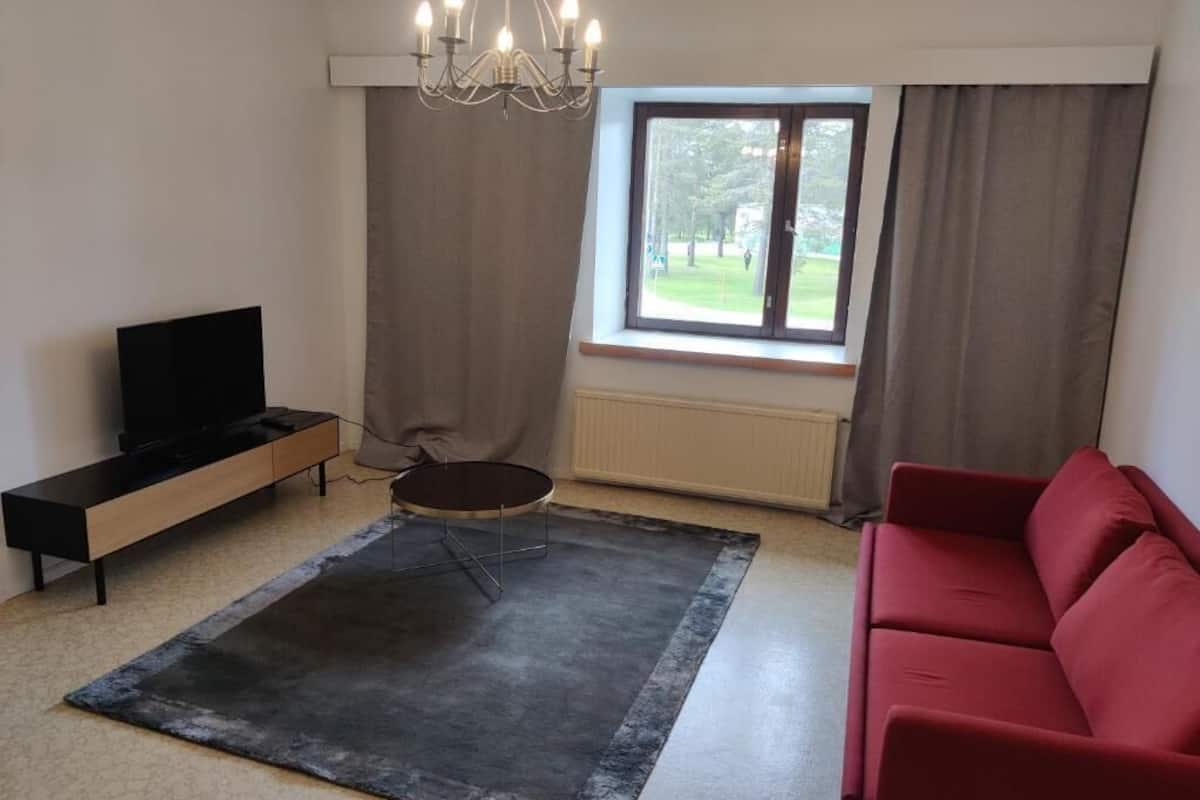 A living room with a red sofa, a black TV stand, a round coffee table, a chandelier, and a window with curtains.