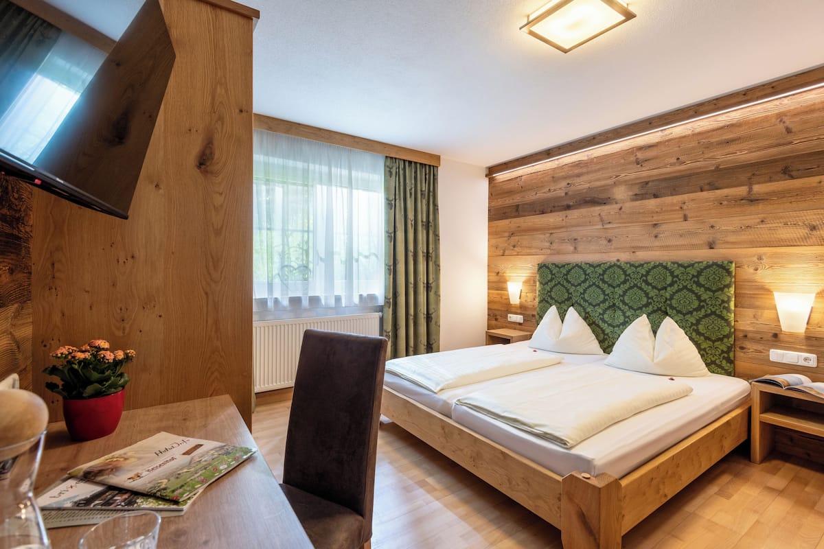 Un dormitorio con cama de madera, un televisor montado en un panel de madera, un escritorio con silla, una maceta roja y una ventana con cortinas de encaje.