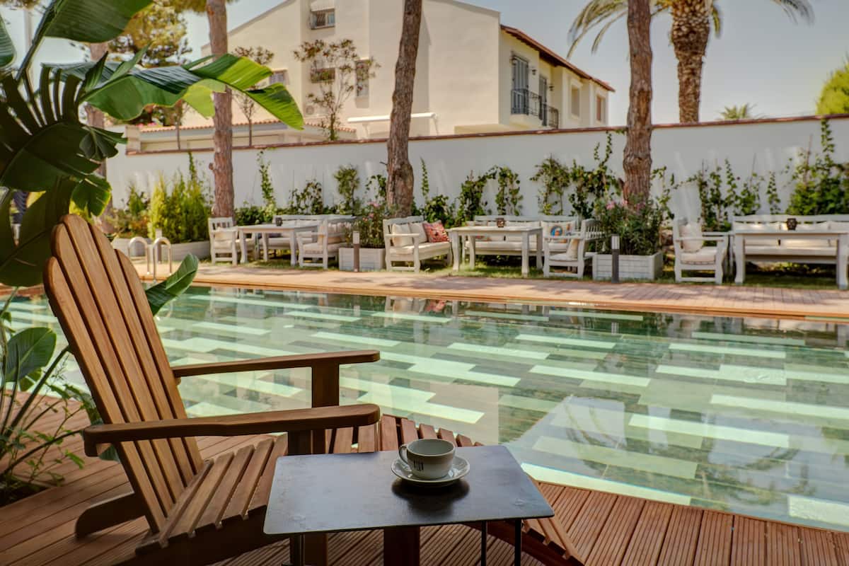 A poolside area with wooden chairs and tables, a glass table, and a view of a building with palm trees.