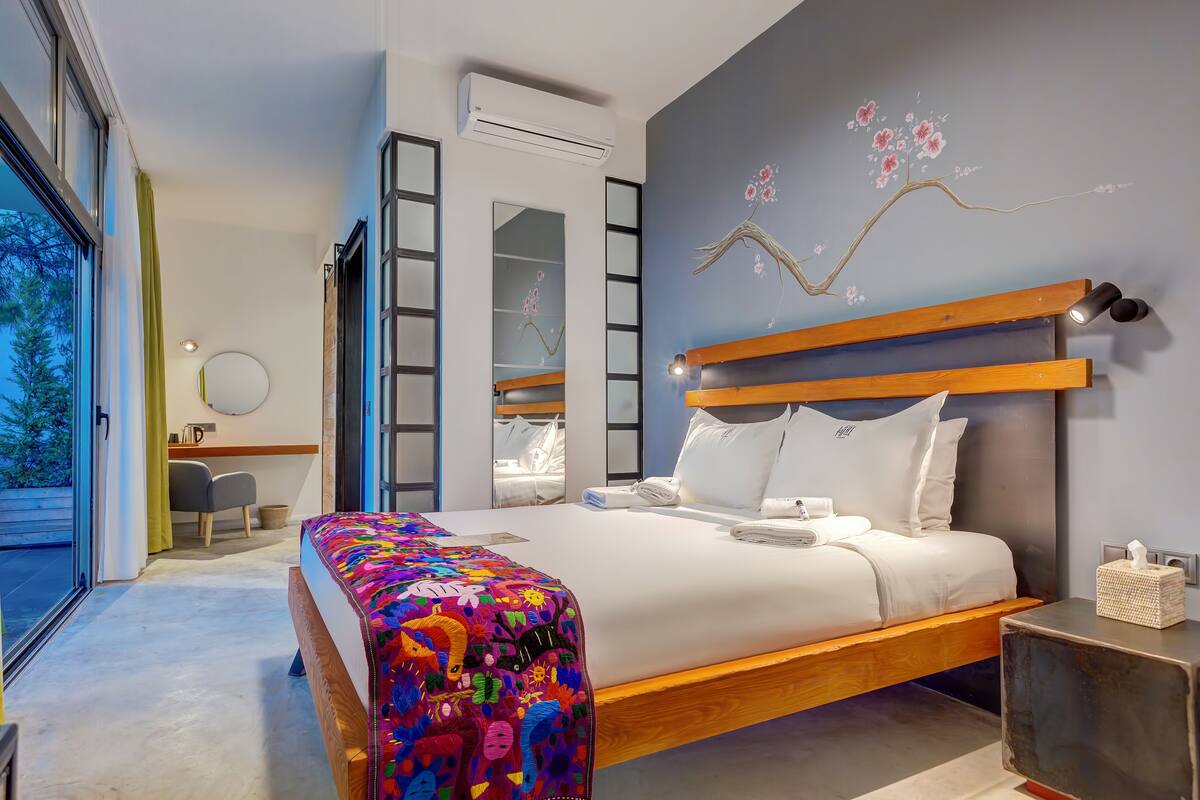 A modern bedroom with a large bed, a wooden headboard, a colorful blanket, a bedside table with a tissue box, and a wall-mounted air conditioner.
