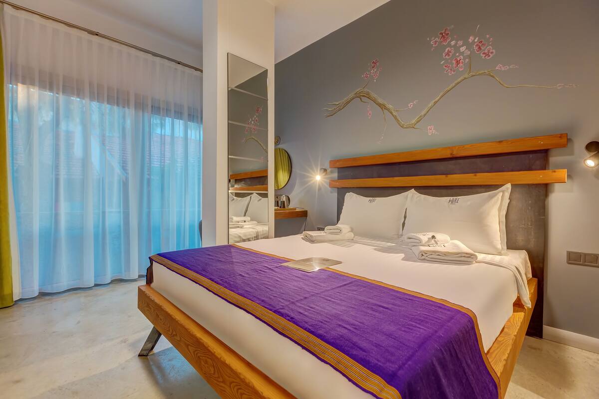 A modern bedroom with a large bed, a wooden headboard, a purple bedspread, and a wall mural of a tree branch with flowers.