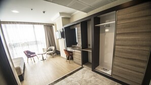 Quarto de hotel moderno com uma grande janela, área de estar com uma cadeira e mesa, cozinha compacta e armário com porta deslizante.