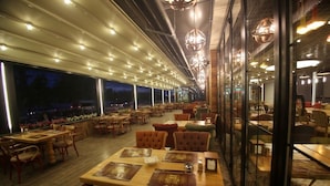 Um restaurante com várias mesas e cadeiras, janelas amplas e luminárias pendentes.