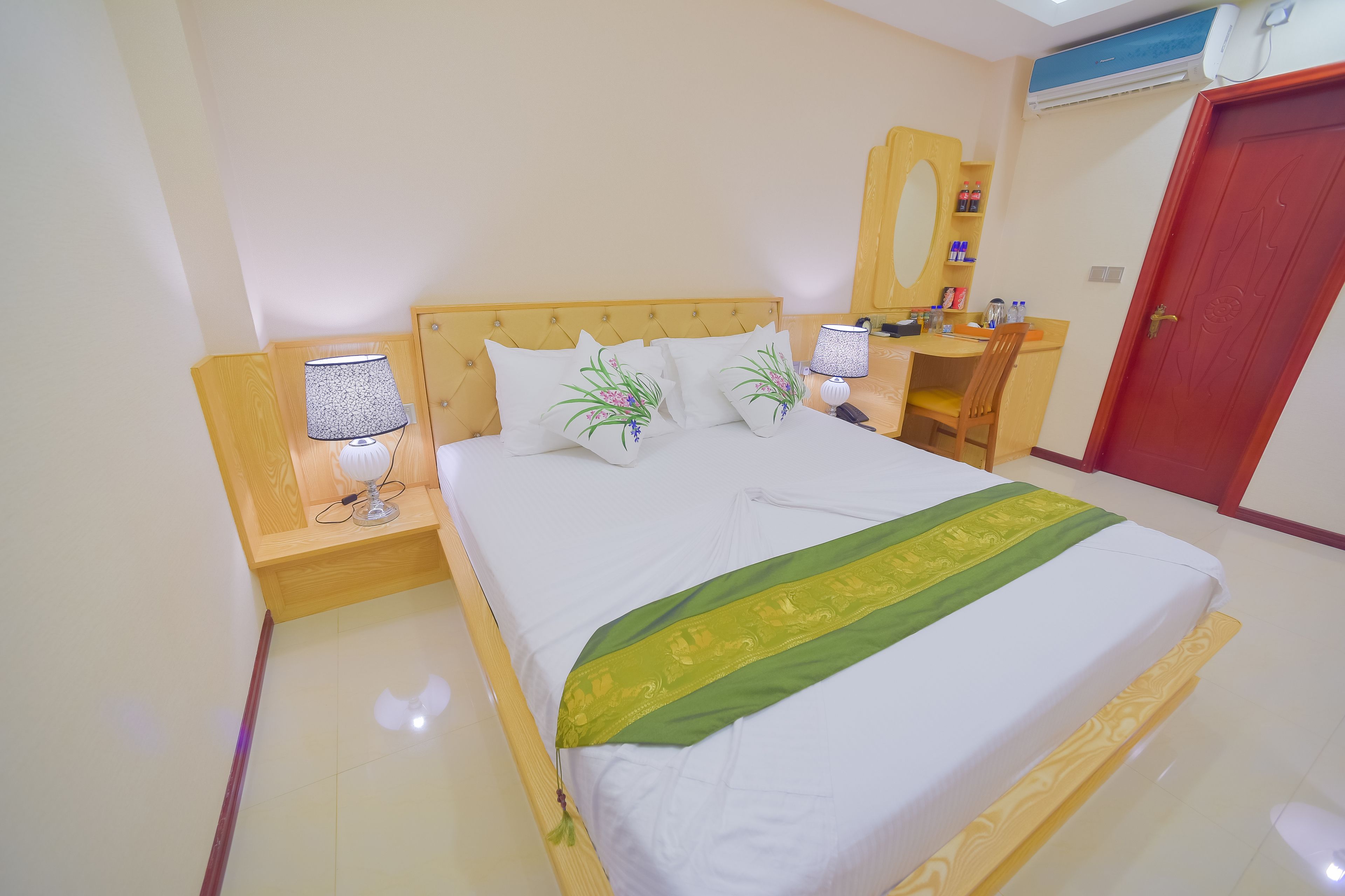 A neatly made bed with a green and white bedspread, two bedside lamps, a wooden nightstand, a mirror, and a red door.