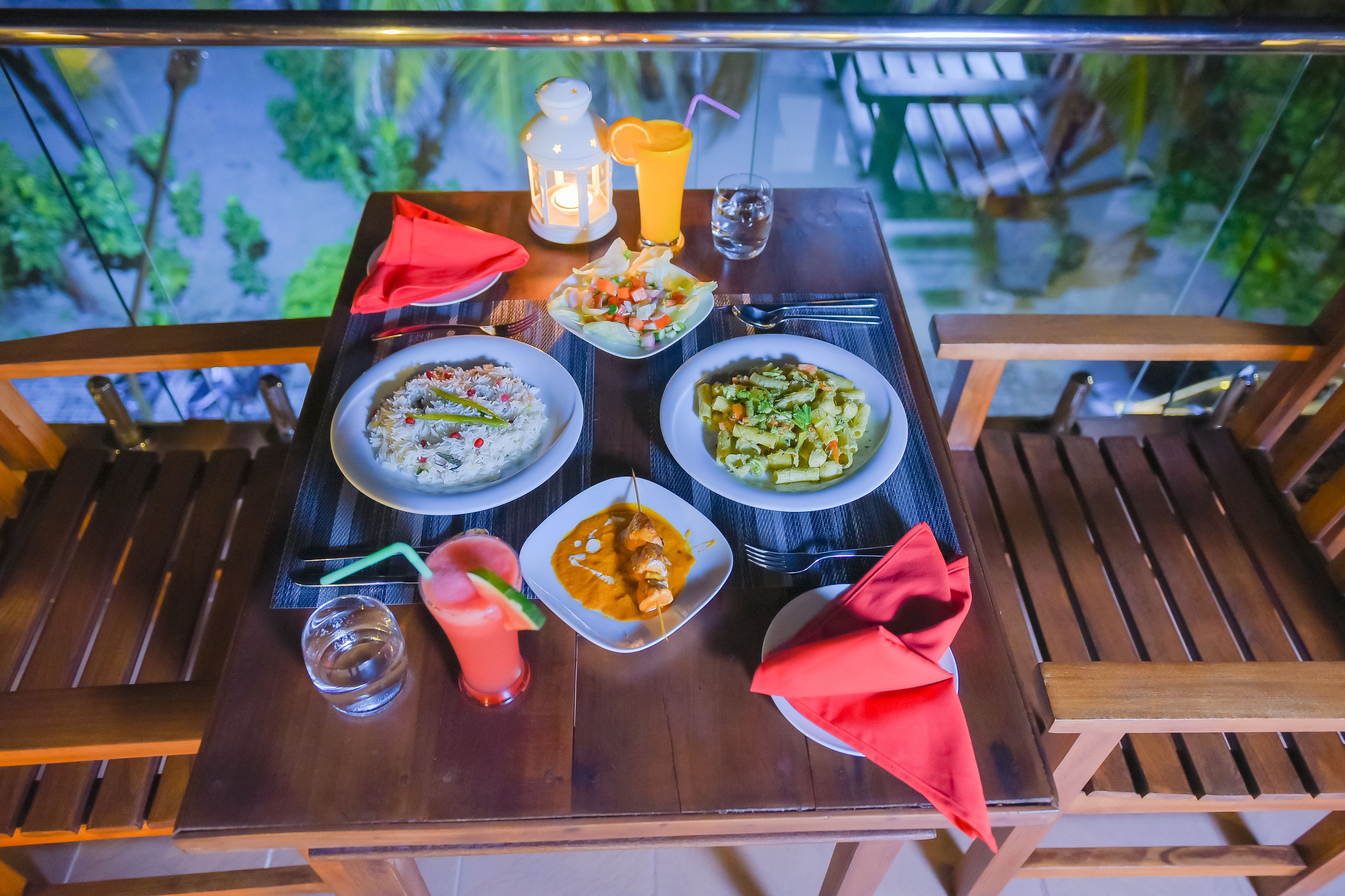 Una mesa de madera con cuatro platos de comida, un farol, una bebida y un mantel individual rojo.