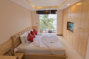 Habitación de hotel con cama, televisor montado en la pared y balcón con vistas a palmeras y al océano.