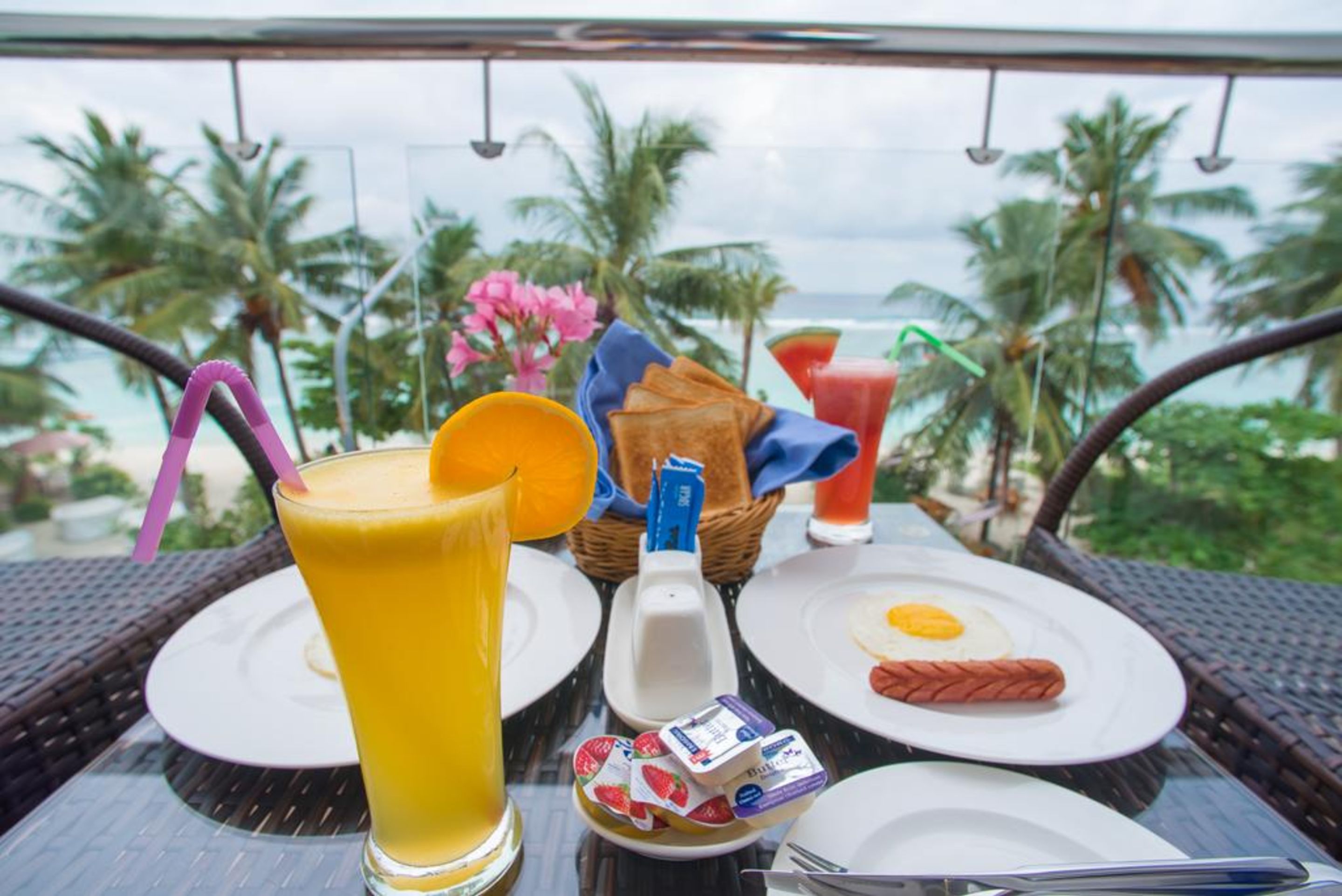 Una mesa de desayuno con un vaso de jugo de naranja, un perro caliente, tostadas y una bebida con pajita, frente a una vista de palmeras y el océano.