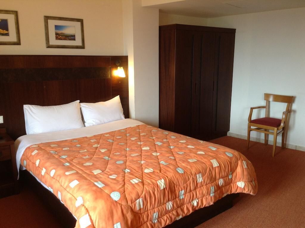 A hotel room with a large bed, a wooden wardrobe, a wooden chair, and two framed pictures on the wall.