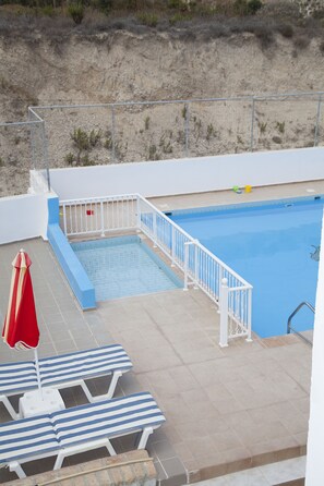 Une piscine avec une balustrade blanche, des marches bleues et un parasol rouge. On y trouve des chaises longues à rayures et une petite poubelle à proximité.