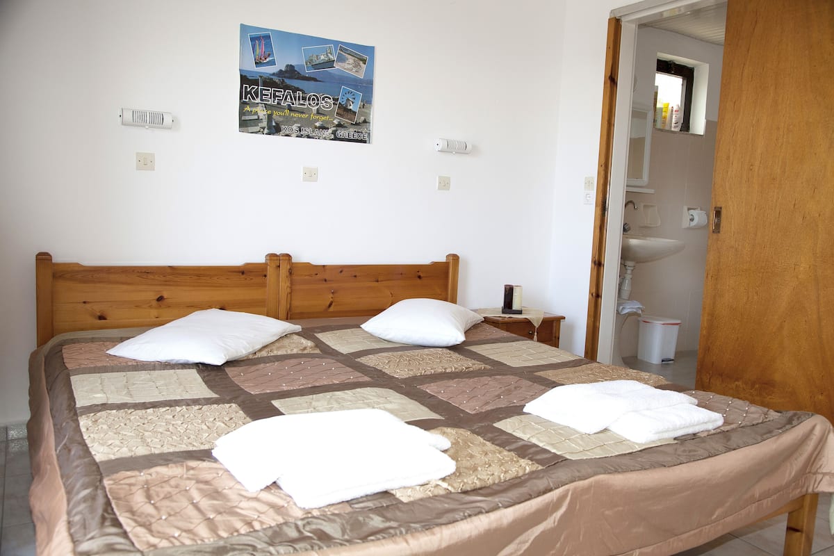 Una cama bien hecha con colcha, almohadas y toallas dobladas. Hay un cartel en la pared y se ve un baño a través de una puerta abierta.