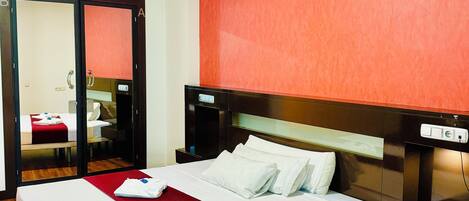 A hotel room with a large bed, a wooden floor, a red accent wall, and a mirror.