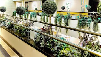 Un espacio interior moderno con barandilla de vidrio, varias plantas en macetas y una pared con letreros numerados.