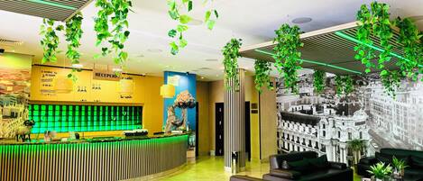 A hotel lobby with a reception desk, seating area, and green plants hanging from the ceiling.