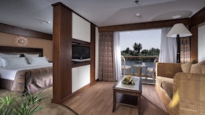 A hotel room with a bed, a sofa, a small table with a fruit bowl, a TV, and a balcony with a view of the water. - Sonesta Star Goddess Nile cruise ship - 4 & 7 nights - From Luxor every Saturday (Luxor)