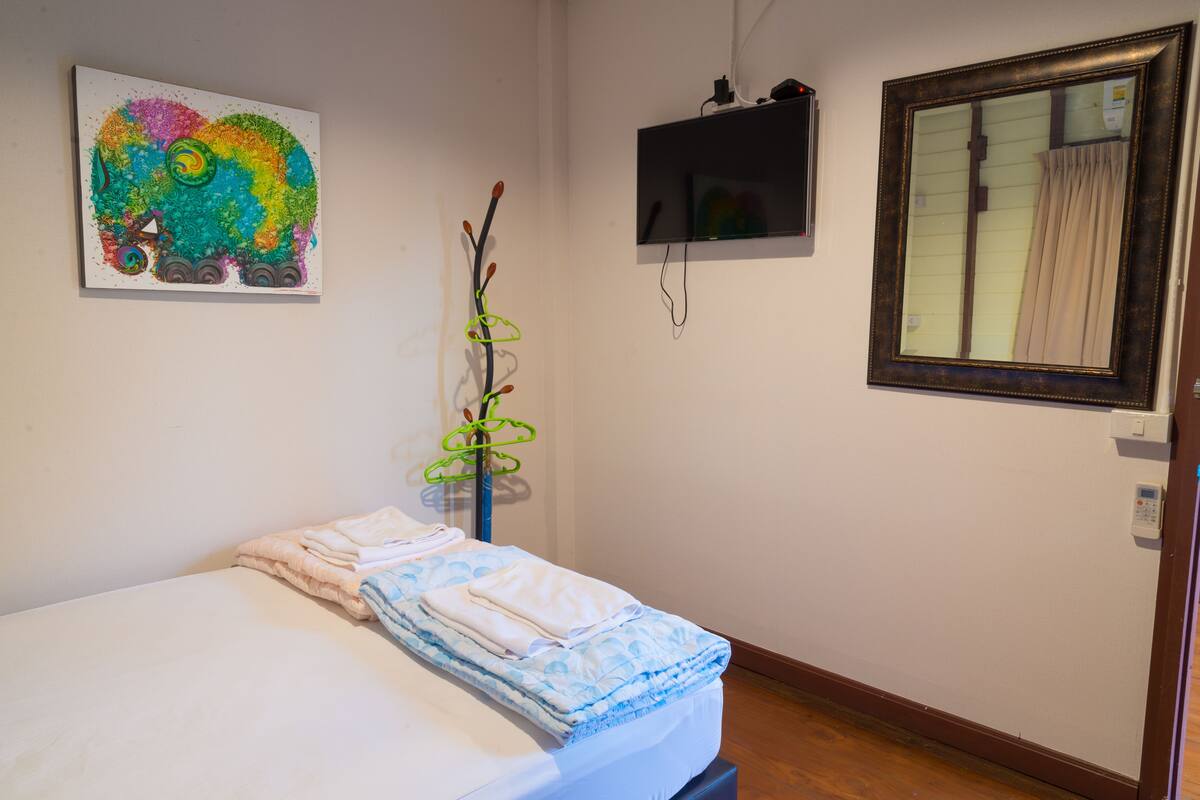Ein ordentlich bezogenes Bett mit einer blauen und weißen Bettdecke, ein farbenfrohes abstraktes Gemälde an der Wand, ein darüber montierter Fernseher und ein Fenster mit Vorhängen.