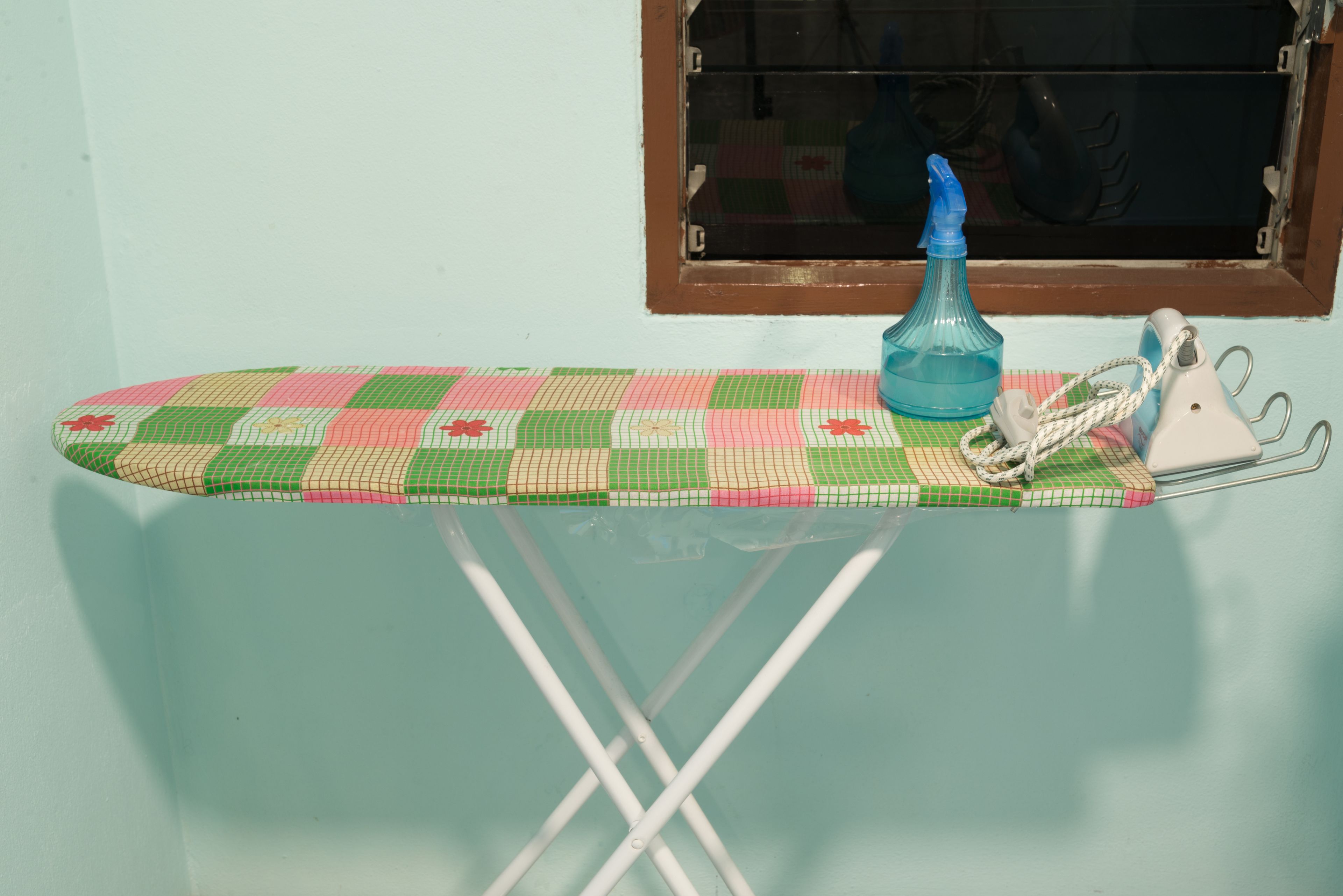 Une planche à repasser recouverte d’un tissu à motifs, un vaporisateur bleu et un fer à repasser blanc avec fil.