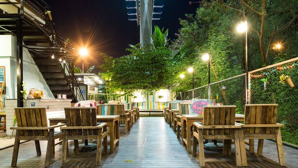 Une terrasse extérieure en bois, dotée de tables et de chaises, entourée de verdure et éclairée par des lampadaires.