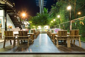 An outdoor wooden deck with tables and chairs, surrounded by greenery and illuminated by street lamps. - NTY Hostel BKK Airport (Bangkok)