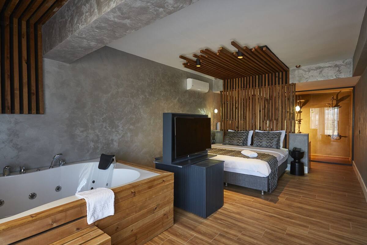 A modern bedroom with a large bed, a TV, a wooden bench, and a bathtub.
