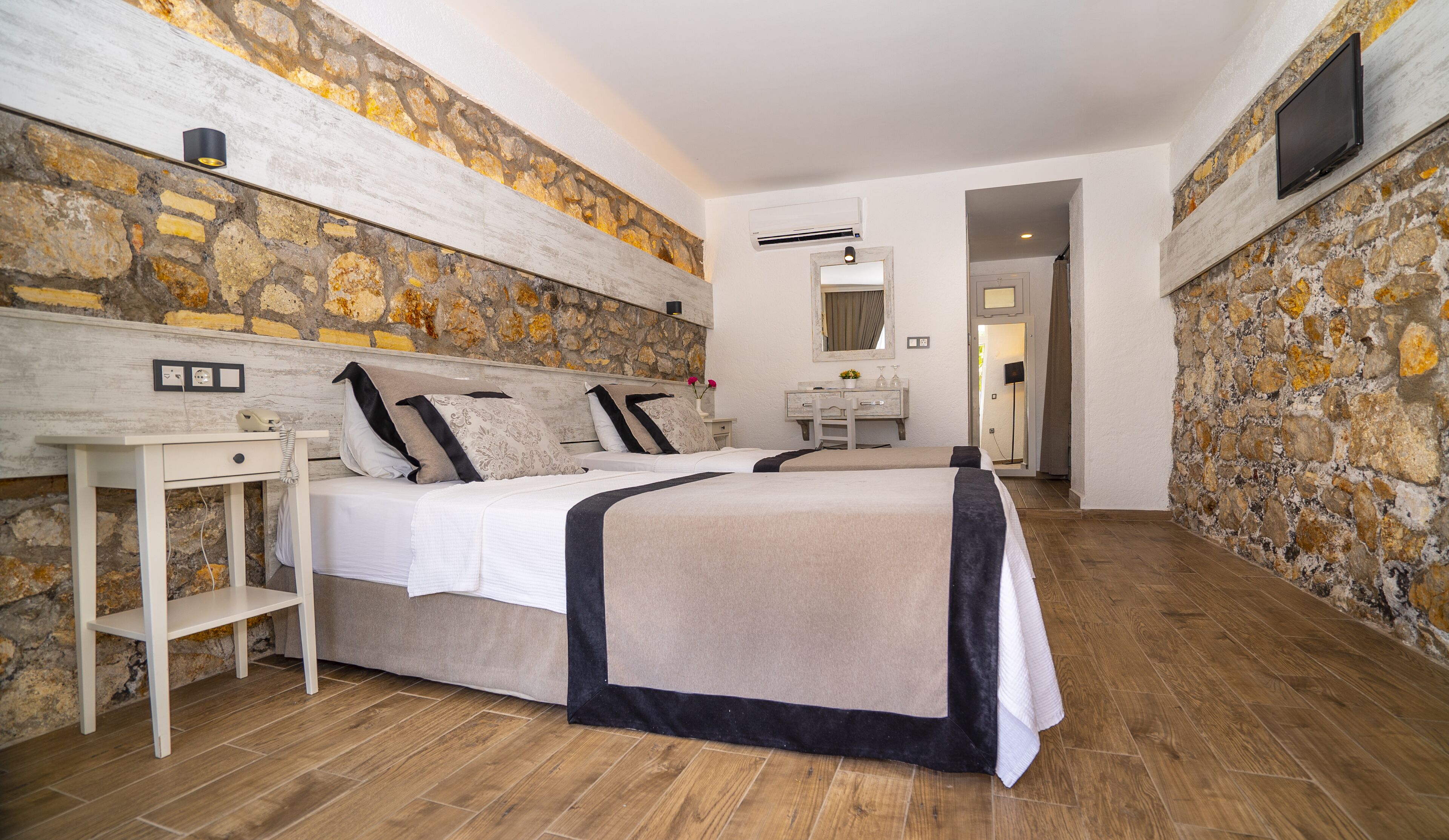 A bedroom with a stone wall, a bed with a white and black bedspread, a bedside table, a desk, and a mirror.
