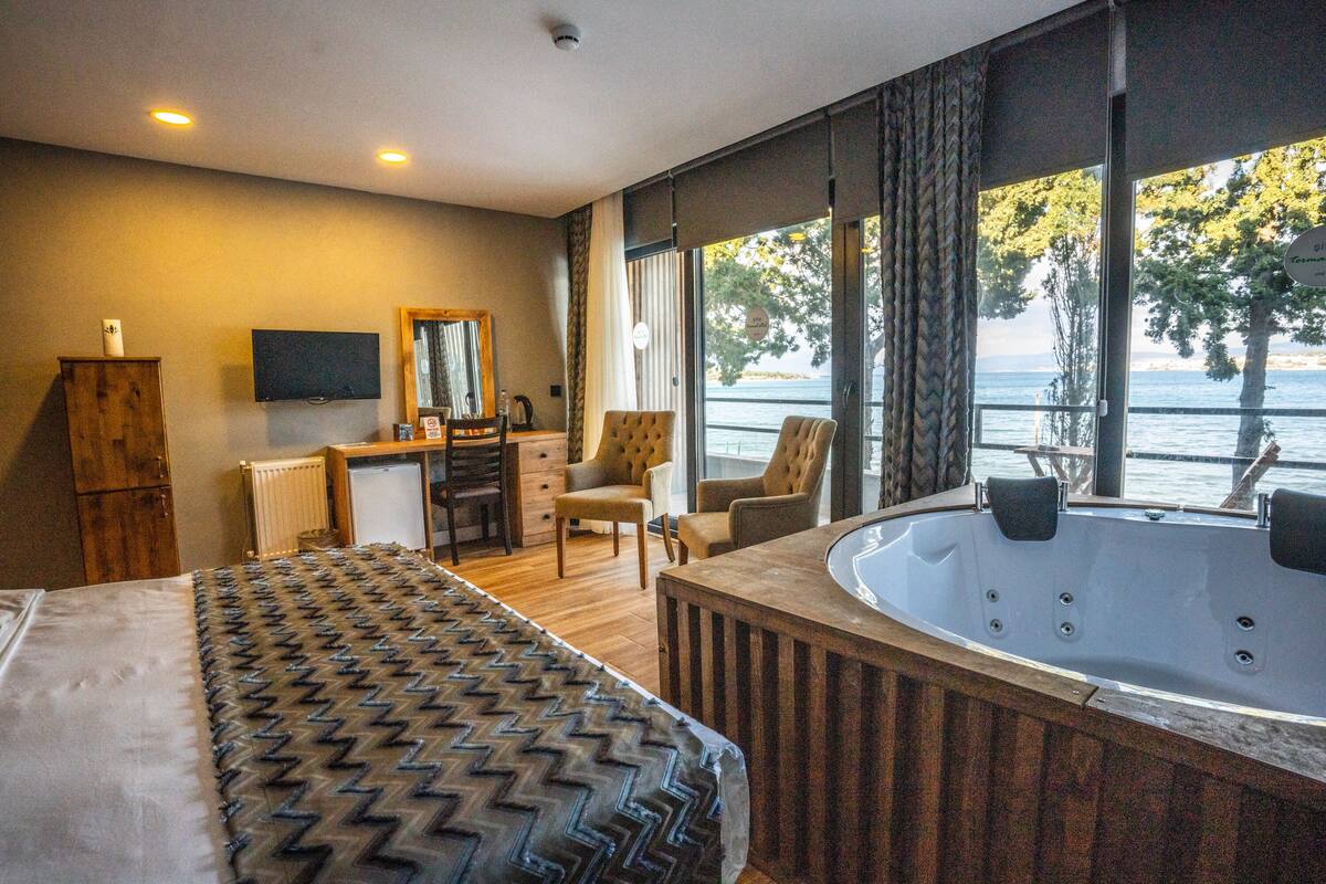 A hotel room with a large bed, a desk, a chair, a television, and a hot tub with a view of the water.