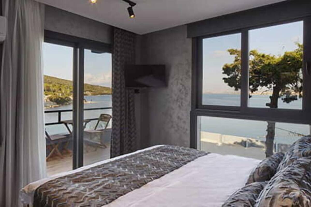 A bedroom with a large bed, a TV mounted on the wall, and a balcony with a view of the sea.